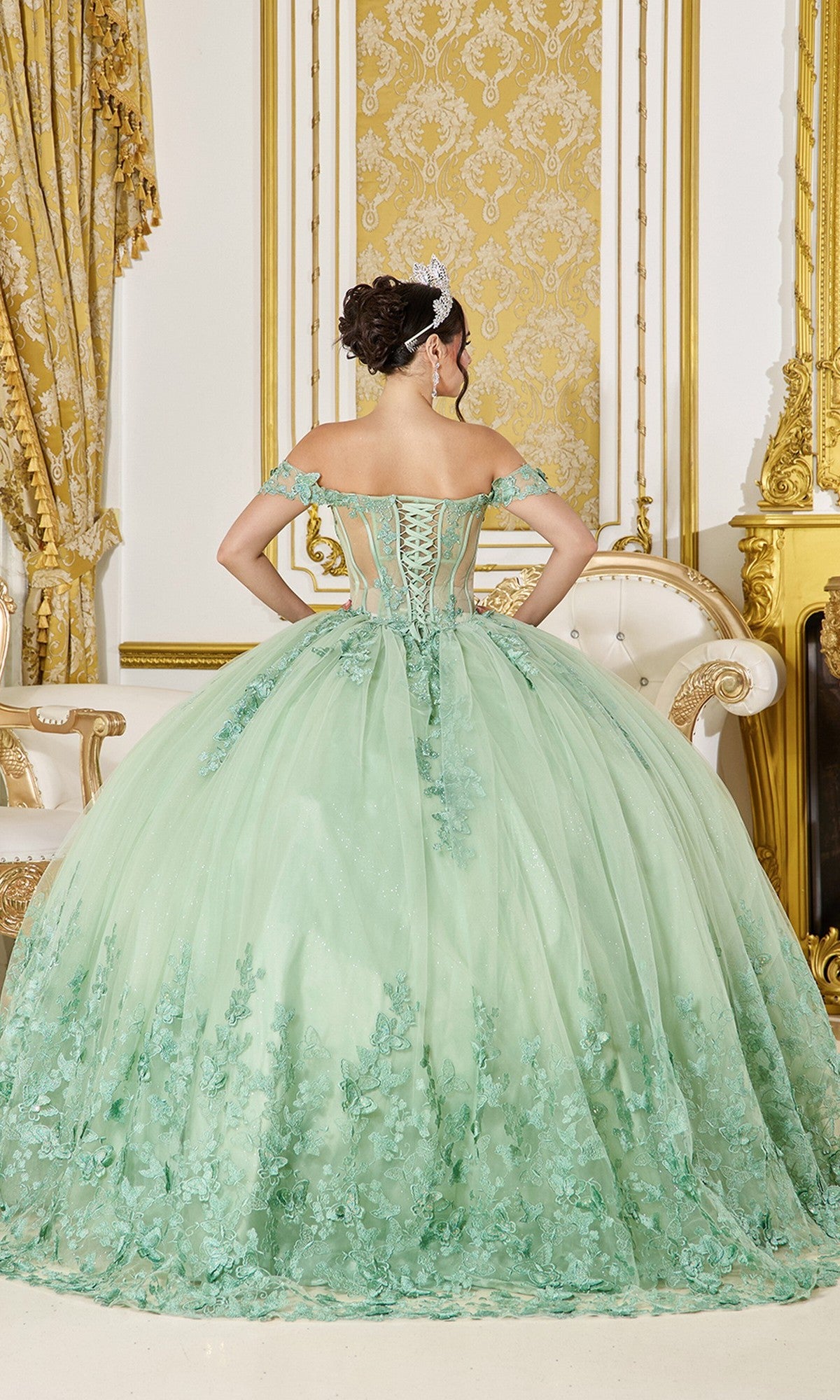 A woman stands with her back to the camera wearing the Sage Green Quinceanera Dress 5065 By Dancing Queen, adorned with 3D butterfly appliqué and floral details, in an ornate room with gold accents.